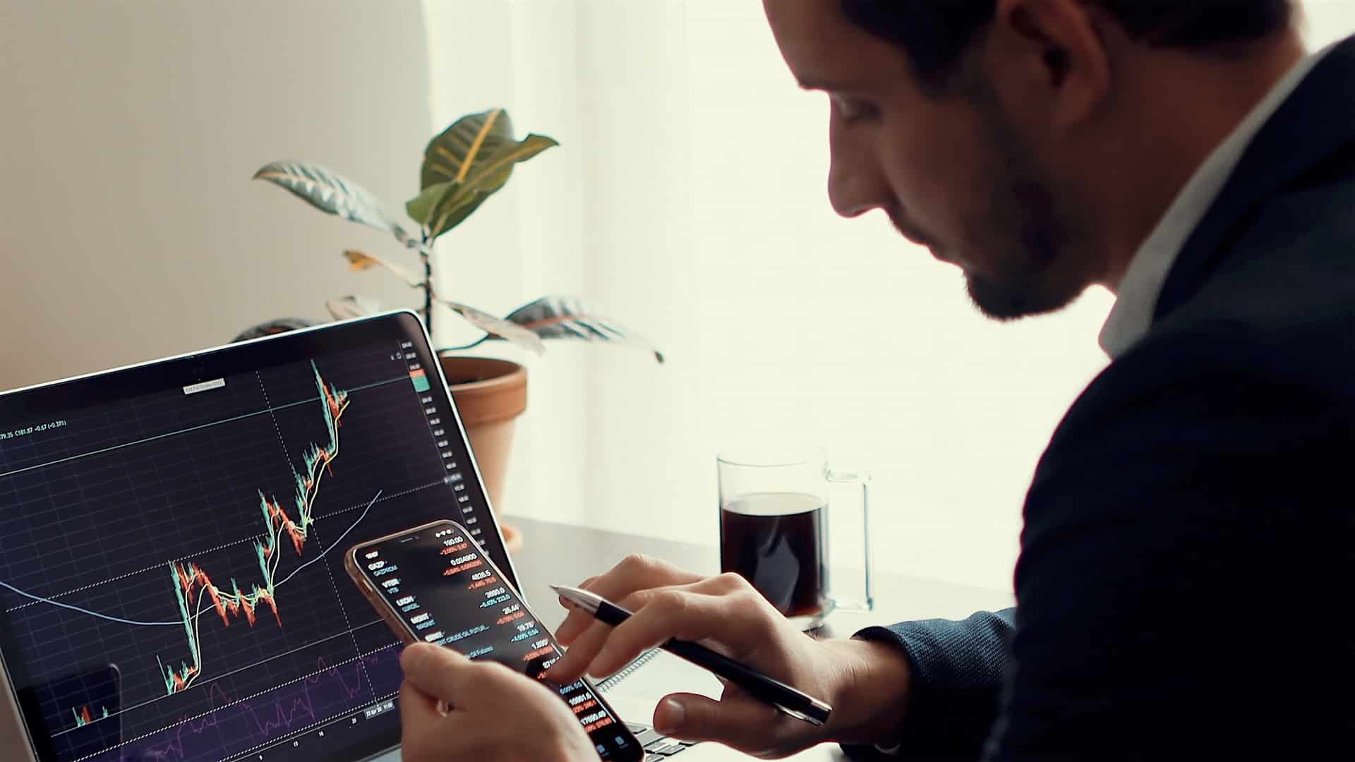 Photo of a man using laptop and a phone app for working in trading.